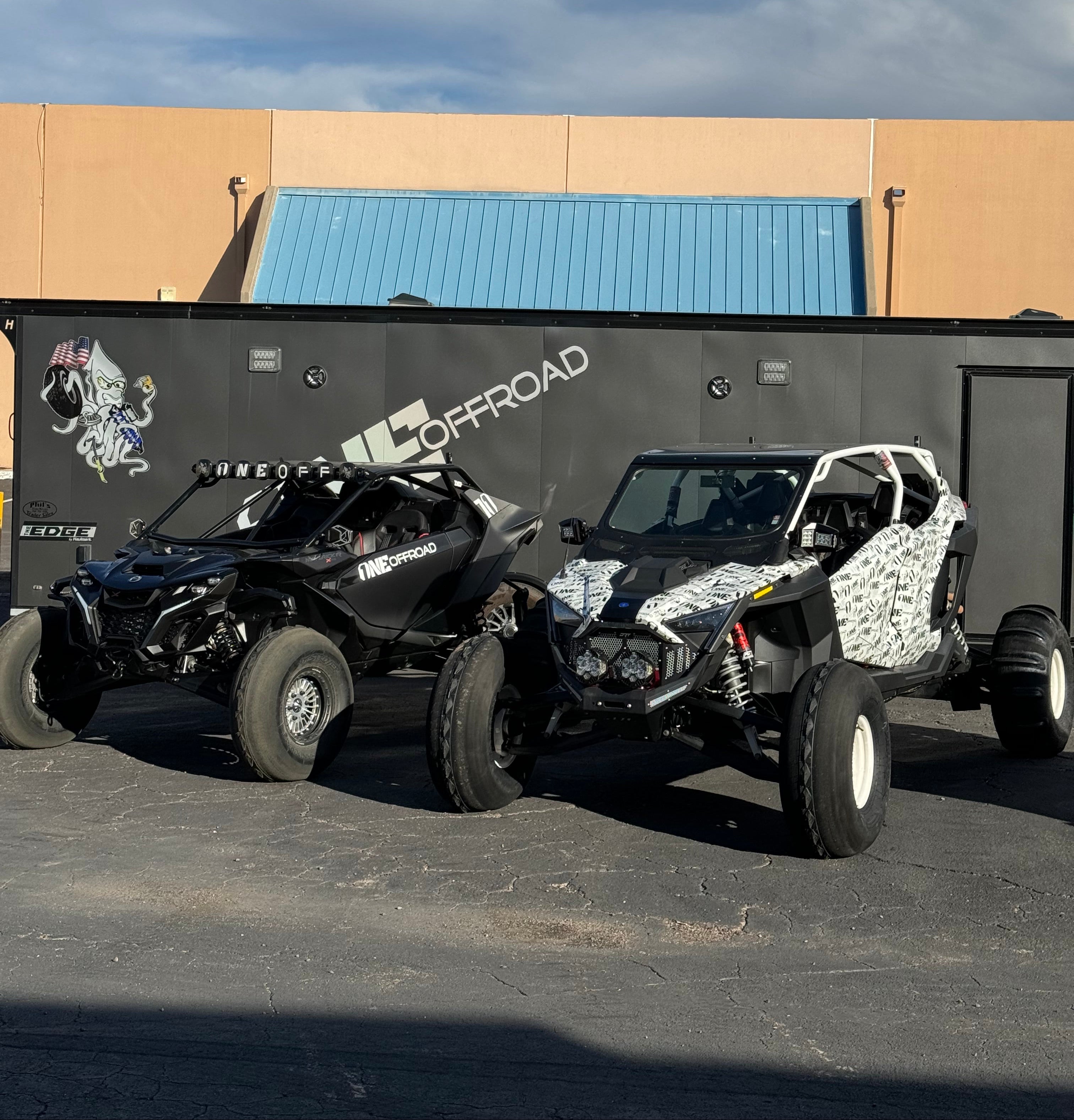 A black off-road dune buggy with white and black patterned fabric on the roll cage, parked outdoors next to a beige building with a blue door under a blue sky.